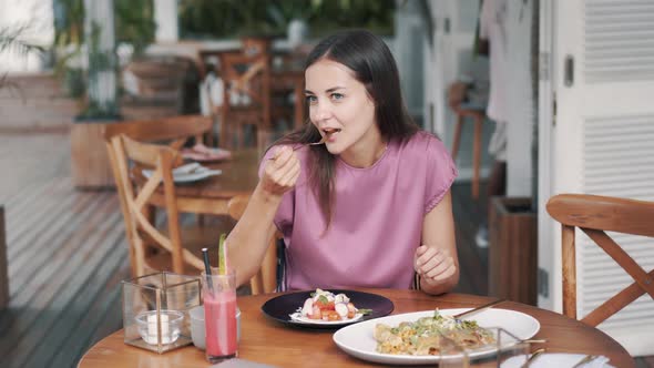 Young Woman Sits at Table in Cafe and Eats Salad of Fresh Vegetables