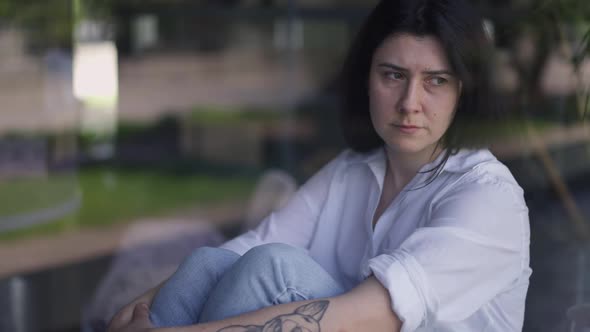 Shooting Though Window of Depressed Caucasian Tattooed Woman Sitting on Windowsill Thinking