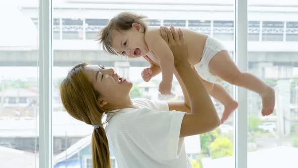 Happy young adult asian mom holding cute adorable infant child girl lifting up standing in bedroom