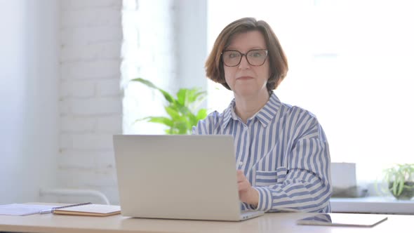 Senior Woman Pointing at Camera While Using Laptop