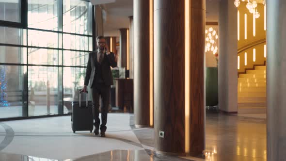 Young Man in Suit Carrying a Suitcase Walking Down the Hotel Lobby and Talking on Mobile Phone