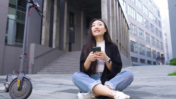 Asian Woman Sitting on Concrete Ground on the City Building Background and Using Her Smartphone