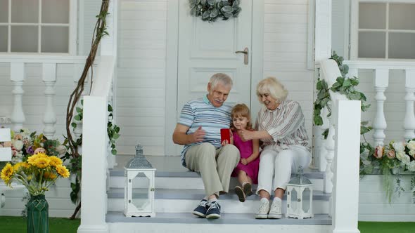 Senior Ouple Sitting with Granddaughter in Porch at Home. Using Mobile Phone for Online Video Call