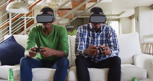 African american teenage twin brothers on couch using vr headsets and playing computer game smiling