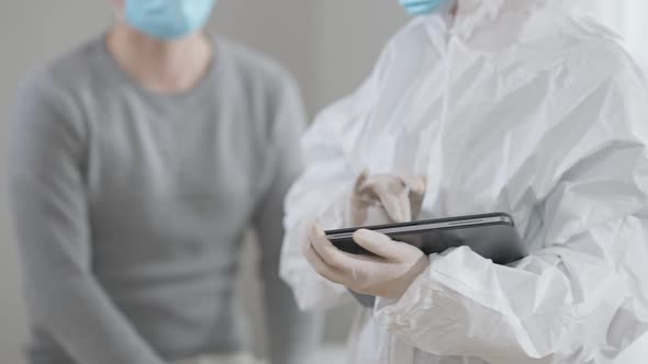 Unrecognizable Doctor in Protective Uniform and Gloves Typing on Tablet As Patient in Coronavirus