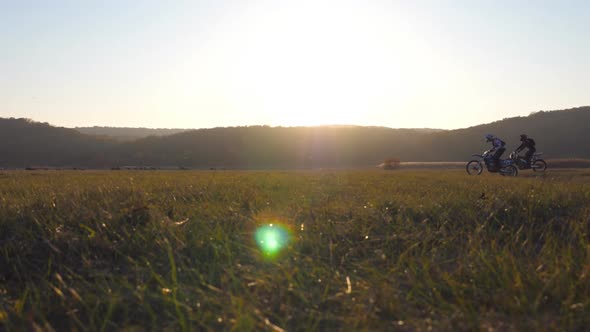 Two Motorcyclists Passing Through Large Field with Beautiful Sunset at Background