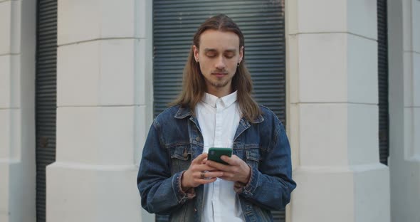 Hipster Young Man with Earrings Looking Aside While Using His Smartphone. Bearded Millennial Guy