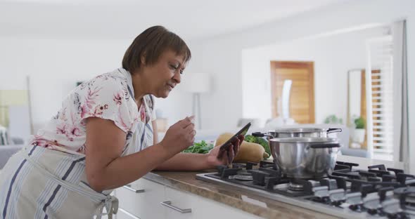 Happy african american senior woman preparing food in kitchen, using tablet and smiling