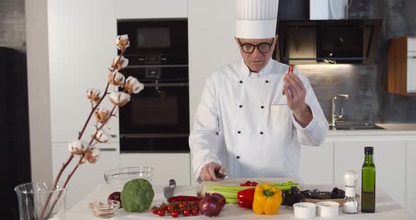 Smiling Aged Chef in Uniform Standing in Kitchen and Cutting Pepper While Filming Himself for Blog