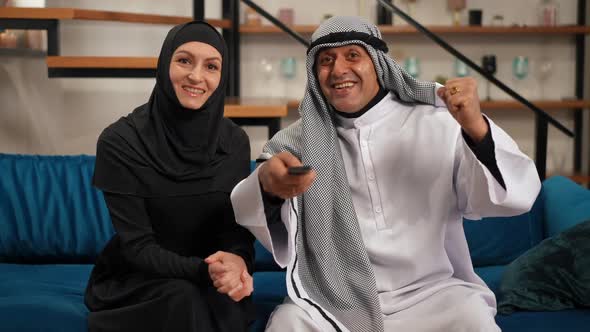 Excited Man in Keffiyeh Sitting on Couch with Woman in Hijab Watching Sports Match on TV Talking