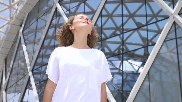 Fashionable Young Woman In Wireless Earphones Against Contemporary Architecture In City