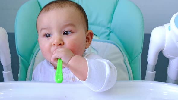 A hungry baby is gnawing on a plastic spoon at the table on a high chair. Teething, whims, itchy gum