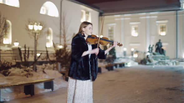 An Attractive Girl is Standing in the Square in a Long white Dress and Playing the Violin