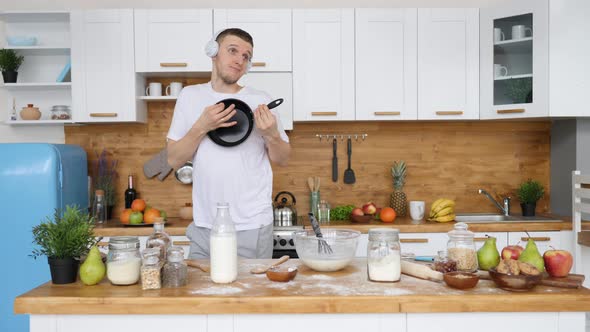Young Handsome Man Playing On Frying Pan In Headphones While Cooking In Kitchen.