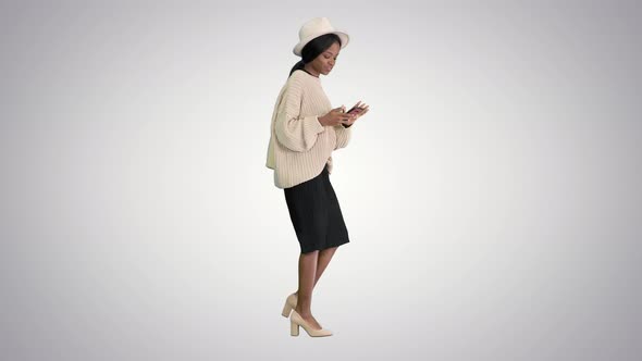 Stylish African American Woman in Knitwear and Hat Using Her Phone While Walking on Gradient