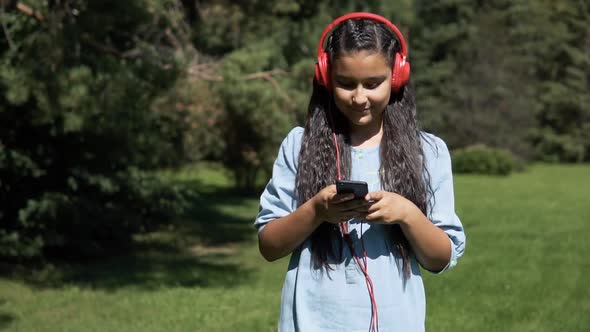 An Attractive Girl with Long Hair Walks Through the Park and Listens to Music in Headphones