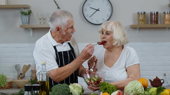 Senior Couple in Kitchen. Grandmother and Grandfather Feeding Each Other with Raw Vegetable Salad