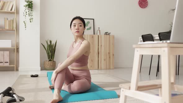 Young Woman Sitting on a Yoga Mat Following an Online Fitness Class on Laptop