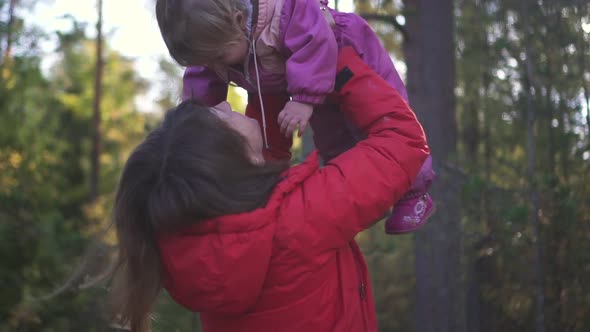 Happy Mom in Jacket Holding Daughter in the Air and Kissing Her in Green Forest