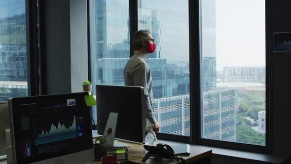Thoughtful caucasian man wearing face mask looking out of window at modern office
