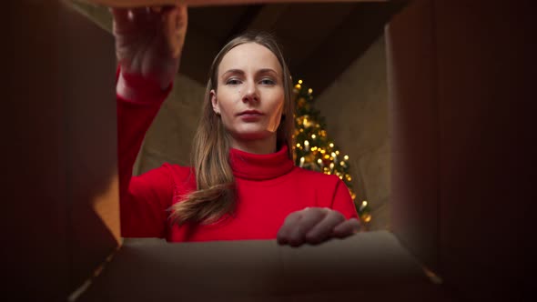 Cheerful Pretty Young Woman in a Red Sweater Opens a Christmas Gift