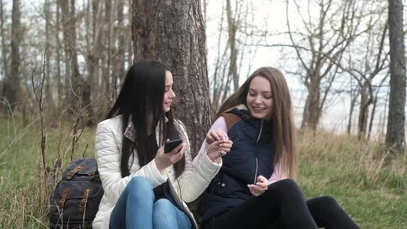 two girls sitting near tree on grass listen to music smartphone with headphones