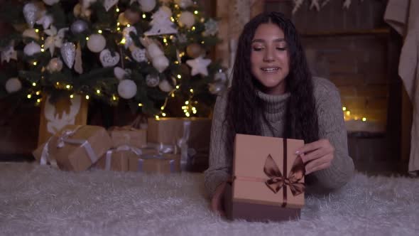 The Girl Opens the Box with a Gift and Rejoices Lying on the Floor Near the Christmas Tree