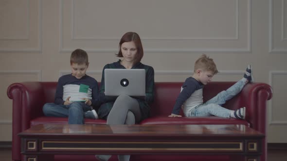 Young 30s Mother with Laptop and Two Boys in the Same Clothes Using Gadgets, Sitting on Couch