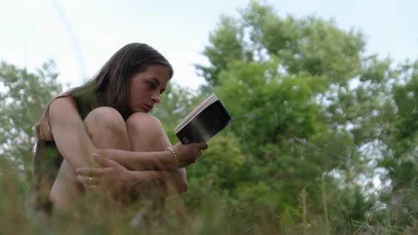 Young Woman Reads a Book Sitting on the Grass in Nature at Summer Day