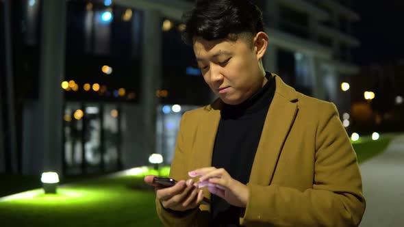 A Young Asian Man Works on a Smartphone in a Park in a City at Night - Closeup