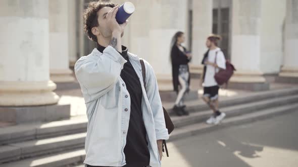 Tattooed Curlyhaired Caucasian Man Drinking Coffee and Smiling at Camera with Blurred Hippie Friends