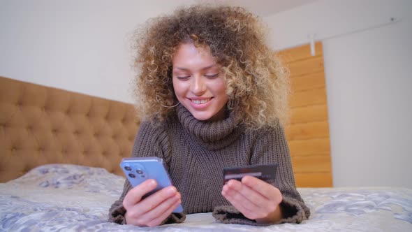 Woman shopping online with smartphone and credit card while lying on bed in bedroom in 4k footage