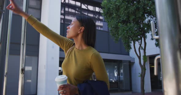 African american woman in street holding coffee and raising hand to stop a taxi