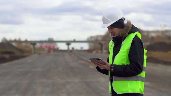 The Engineer Looks at the Construction Plan on the Tablet, on the Background of the Construction