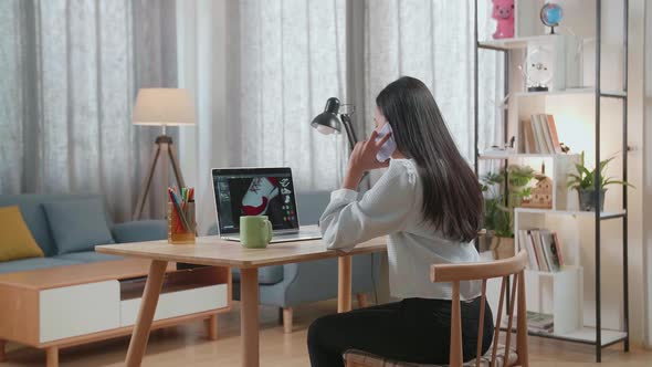 Asian Female Footwear Designer Talking On Smartphone While Designing Shoe On A Laptop At Home