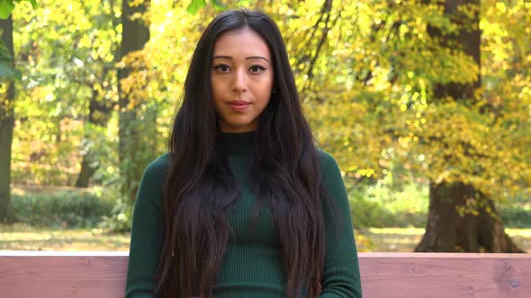A Young Asian Woman Sits on A Bench in A Park and Looks Seriously at The Camera