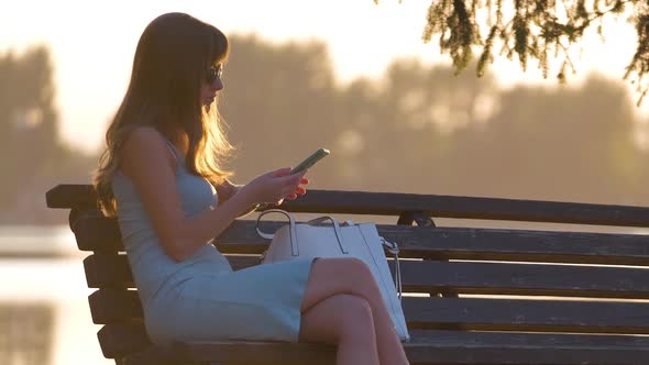 Young Woman Sitting on Park Bench Browsing Her Cellphone Outdoors on Warm Summer Evening