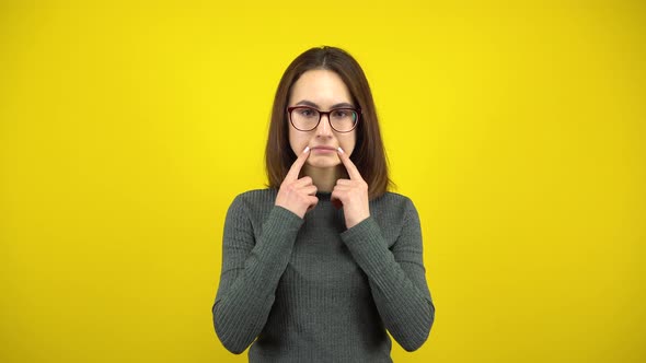 A Young Woman Makes a Sad Smile on Her Face with Her Fingers on a Yellow Background. Woman with