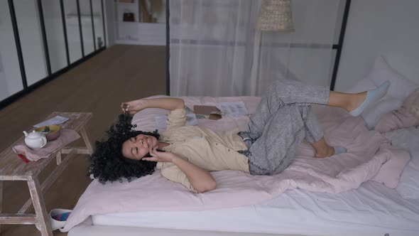 Side View Wide Shot of Charming African American Smiling Woman Lying on Bed Talking on the Phone