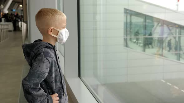 A Little Boy in a Mask Stands Near a Large Window in the Airport Waiting Room