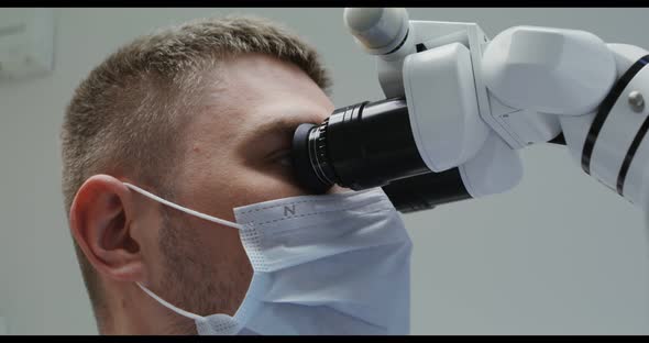 A Male Doctor or Scientist in a Medical Mask Looks Through a Microscope