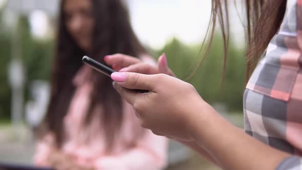 Two Young Girls Sit in a Park Near a Fountain Using a Smartphone and a Tablet in Sunny Weather