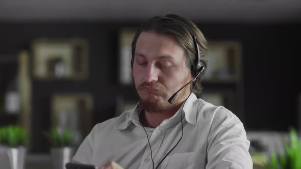 Young Manager in Headphones Is Working in Office Looking in Smartphone