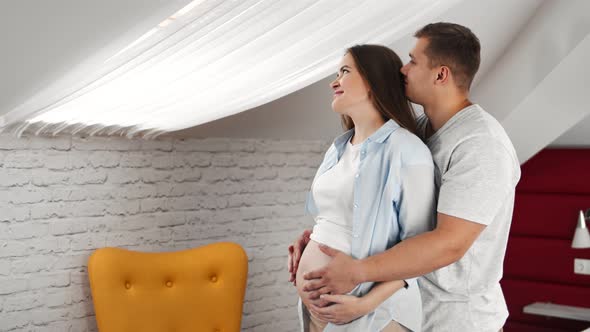 Happiness Young Pregnant Couple Hugging and Kissing Relaxing at Home Looking Out Window