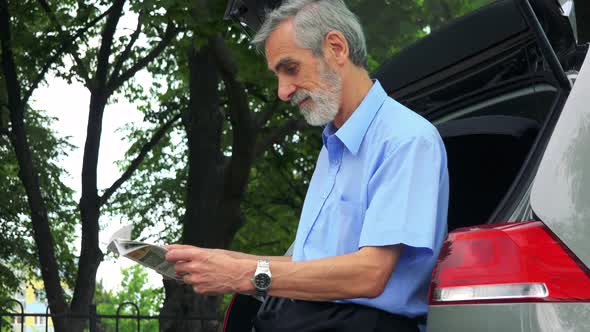 Senior Man Sits in the Trunk and Reads a Newspaper - Trees in the Background