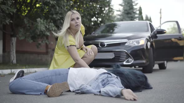 Stressed Young Blond Woman Checking Pulse of Body Hit By Car Outdoors. Portrait of Frustrated Young