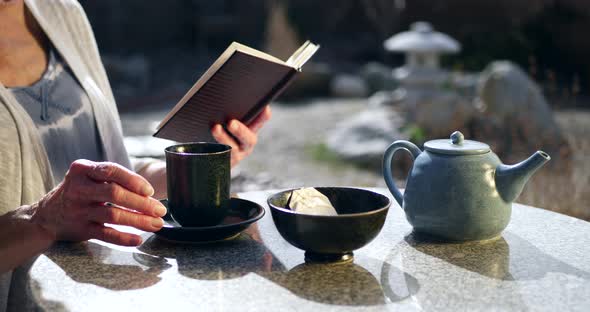 An elderly woman reading a book and drinking a cup of herbal tea with a bowl and a teapot in a Japan