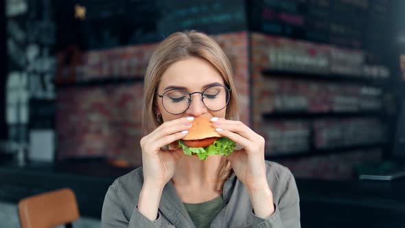 Portrait of Pleasant Smiling Young Woman in Glasses Biting Appetizing Fresh Burger Medium Closeup