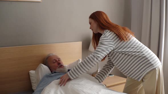 Loving Young Granddaughter Covering Sick Grayhaired Bearded Grandfather with Blanket Lying on Bed at