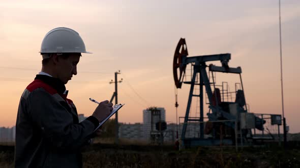 Man in Overalls Fills Paper with a Pen Against the Background of an Oil Pump at Sunset